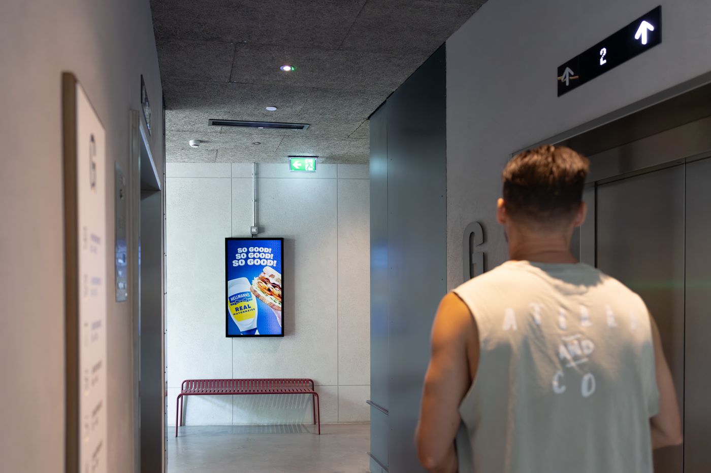 A resident walking towards the lifts in a concrete-and-glass lobby, passing an Abode screen running a live campaign.