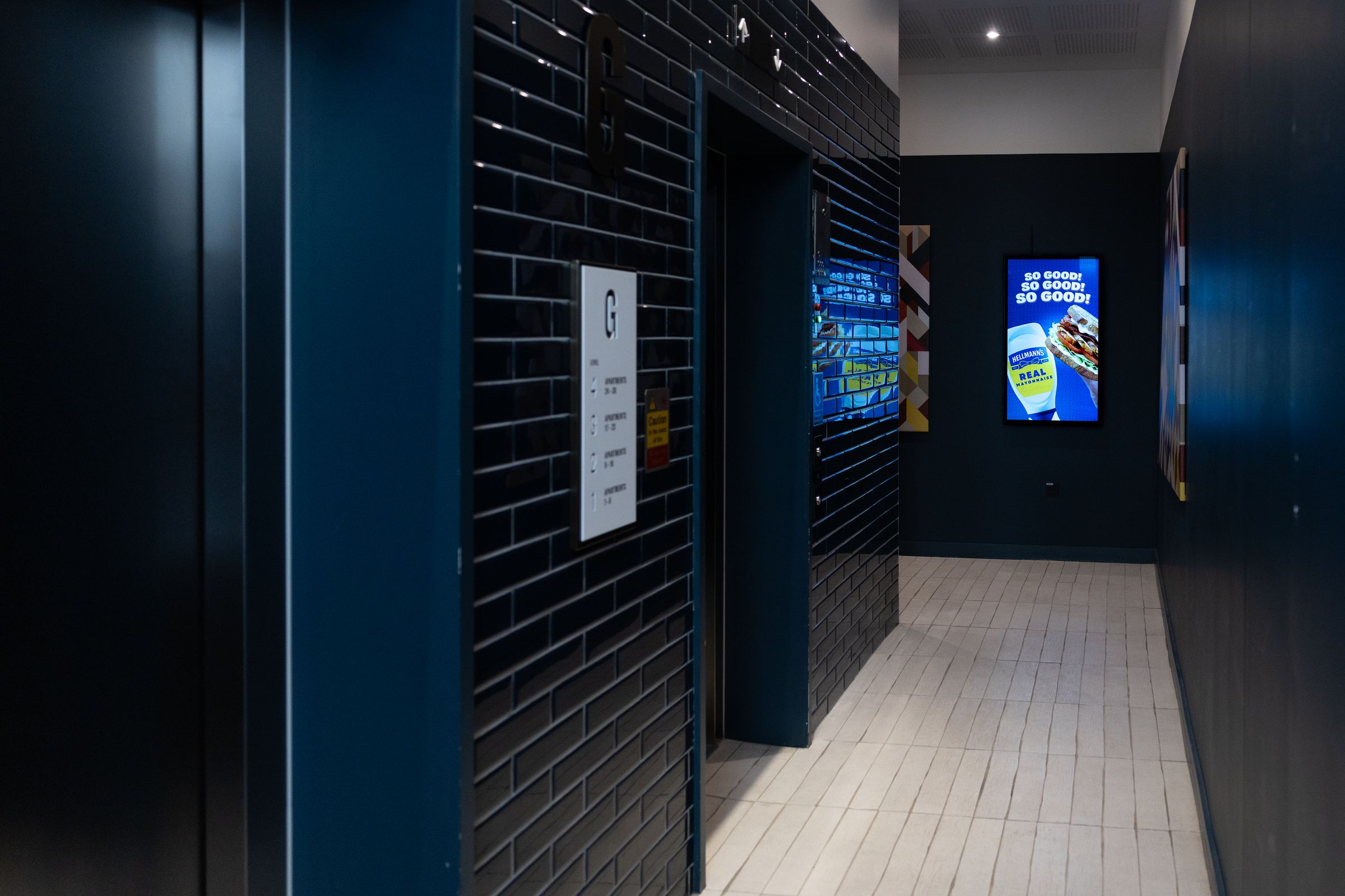 A navy-tiled lift lobby at dusk, an Abode screen glowing on the far wall showing a live Hellmann's campaign.
